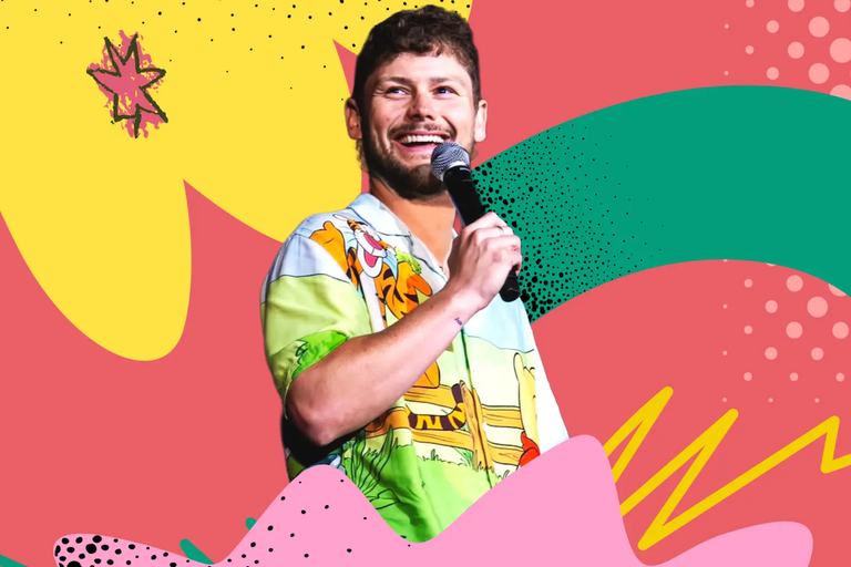 Comedian Drew Lynch smiling on stage into a microphone in front of a colorful pop-art background.